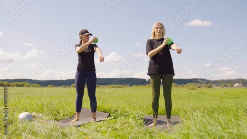 Two young women in sportswear doing myofascial release massage on their forearms and shoulders with a small green ball, standing on yoga mats in a beautiful green field on a sunny day