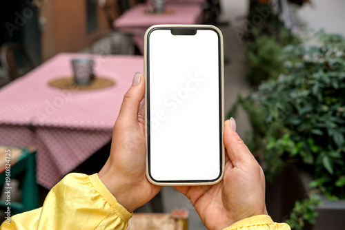 Smartphone mock up held by person wearing  in outdoor cafe setting with tables and greenery in the background, smart phone with white blank screen mobile app design and advertising, online marketing