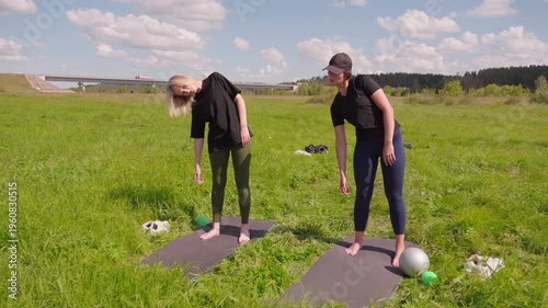 Two athletic women in sportswear doing stretching exercises together on yoga mats in a beautiful green field, enjoying an outdoor fitness workout on a sunny summer day