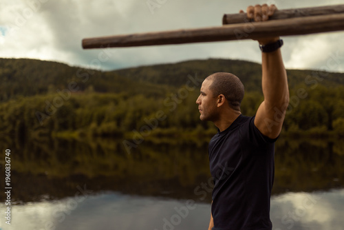 Athletic man in profile holding wooden oars by a calm river. Muscular male athlete looking at distant forested hills under a cloudy sky. Fitness, rowing and outdoor adventure in a scenic landscape.