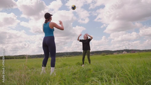 Two athletic young women enjoying a sunny day, playing volleyball in a lush green meadow under a beautiful cloudy sky, embracing an active and healthy lifestyle with outdoor sports and recreation