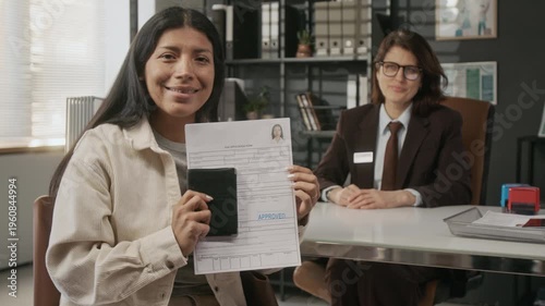 Medium portrait of smiling mature Hispanic woman holding approved visa paperwork after interview with Caucasian female consular officer at embassy, looking at camera with happiness