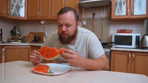 A hungry white chubby man with a beard sits at the table in the kitchen and greedily eats a large piece of ripe red watermelon, enjoying fresh and juicy summer fruits as a healthy snack