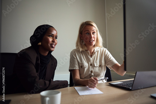 Smiling businesswoman sitting with female colleague explaining on computer at office desk