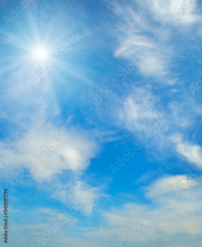 Beautiful vertical shot of a bright shining sun with long rays in a clear blue sky with soft white wispy clouds, representing a peaceful sunny day and high-altitude meteorological weather.