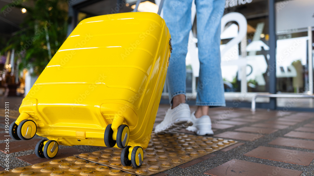 Fototapeta premium Close-up of Asian woman traveler pulling a vibrant yellow suitcase at the airport entrance. Concept of travel, summer vacation, and modern lifestyle with copy space.