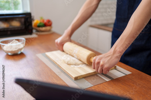 Person's hands using a wooden rolling pin on dough on a wooden board at kitchen counter, following an online recipe