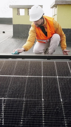 Worker in safety helmet and high-visibility vest cleaning solar panels on a rooftop to ensure efficiency and maintenance of renewable energy system. Vertical video.