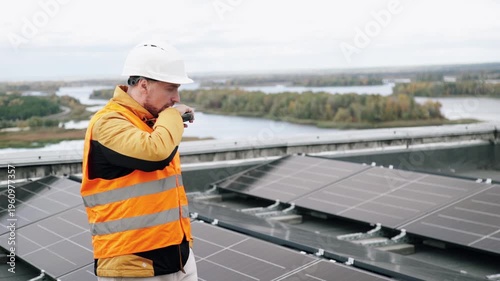 A construction worker wearing a safety helmet and reflective vest inspects the installation of solar panels on a roof, using a walkie-talkie to communicate with his supervisor.
