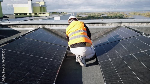An engineer wearing a protective vest and hard hat installs solar panels on a roof using a power drill. Renewable energy technology and sustainable construction concepts.