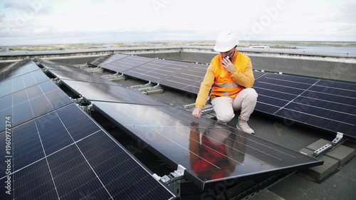 A construction worker wearing a safety helmet and reflective vest inspects the installation of solar panels on a roof, using a walkie-talkie to communicate with his supervisor.