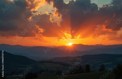 Mountain landscape at sunset. Sun rays burst through clouds illuminating valley below. Orange sky, silhouettes of hills and villages. Calm evening scene.