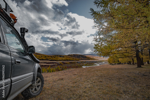 Off-road 4x4 vehicle with snorkel parked on a hill overlooking a scenic autumn valley with a river. Expedition and adventure travel concept under a cloudy sky.