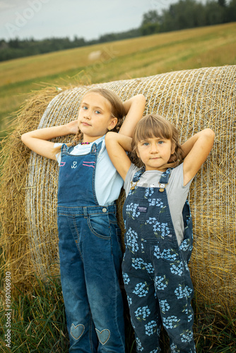 Two cheerful village girls in overalls are standing, leaning on a haystack.