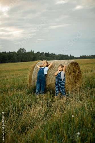 Two cheerful village girls in overalls are standing, leaning on a haystack.