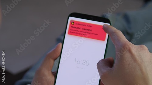 User receives an urgent Tornado Warning emergency alert on a smartphone. A finger taps the red notification. Perfect for disaster preparedness, safety training, and weather-related content. High