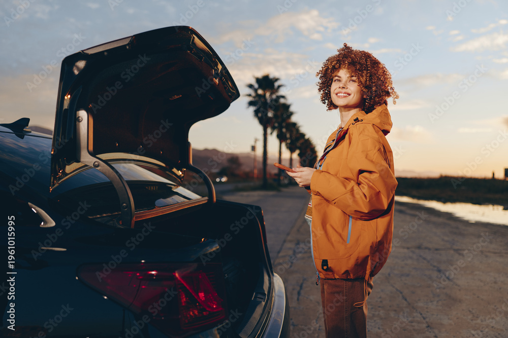 Fototapeta premium Smiling woman with curly hair wearing an orange jacket stands near an open car trunk holding a smartphone on a sunny road lined with palm trees. Lifestyle and travel concept.