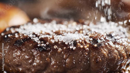 Fine salt crystals being sprinkled over a dark grilled burger patty in slow motion