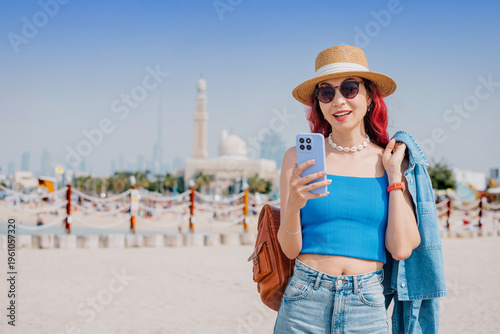 Young woman smiling, wearing a straw hat and sunglasses, holding a phone on a sandy beach with the Dubai city skyline in background