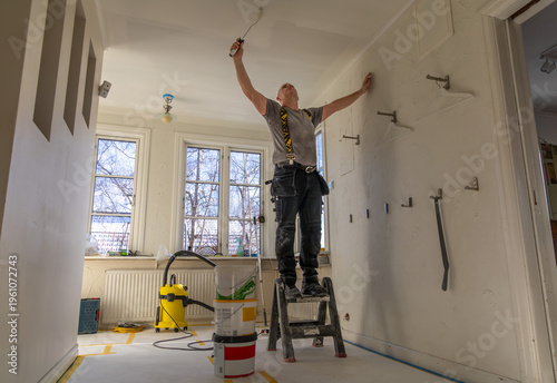 Painter on a step ladder rolling white paint onto a ceiling during renovation.