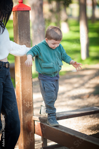 A young boy skillfully walks across a wooden beam in a vibrant park. With sunlight filtering through the trees, he enjoys a playful moment, guided by a caring adult
