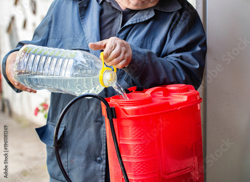 Older man pours solution from plastic bottle into red garden sprayer preparing for pest control treatment of plants in garden