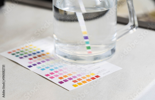 Water quality test strip measurement of parameters in glass pitcher with clear liquid, comparing results with color chart for accurate analysis of water composition.