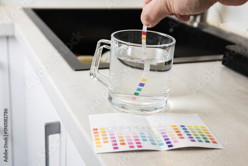 Hand holds water test strip dipping into glass mug with water on kitchen counter next to color chart for measurement of water quality parameters, home water testing process