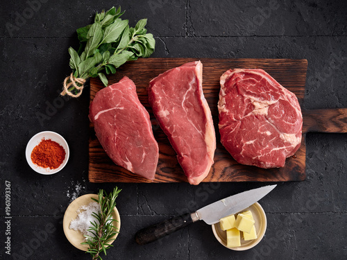 Various cuts of raw beef arranged on a wooden cutting board with herbs and seasoning in a kitchen setting