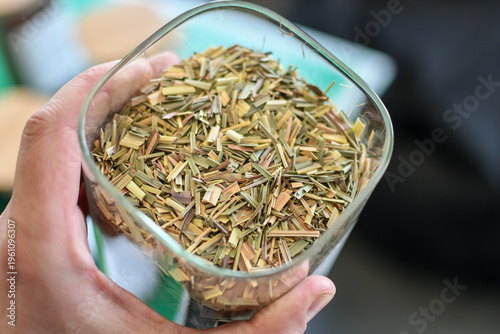 Hand holding dried chopped lemongrass in glass container at herbal processing facility