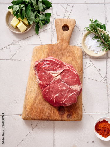 Raw rib-eye steak on wooden cutting board with spices and herbs ready for cooking in kitchen setting