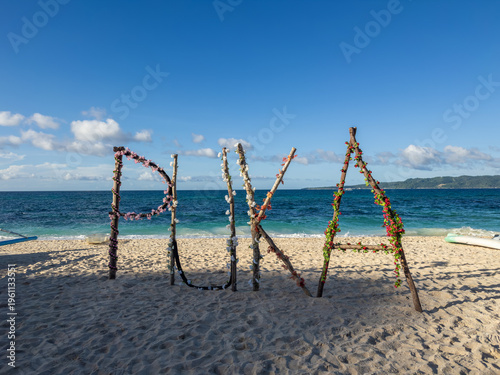 Bild auf Leinwand The iconic 'PUKA' sign, adorned with vibrant blossoms on driftwood, stands on the pristine sands of Puka Beach in Boracay, Philippines