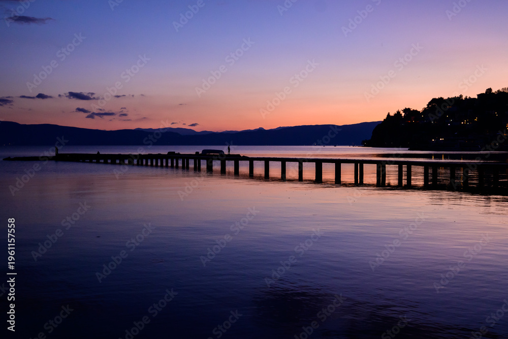 Obraz premium A long wooden pier stretches into the glassy Lake Ohrid, reflecting the stunning purple and orange twilight sky. Silhouettes of people walk along the structure.