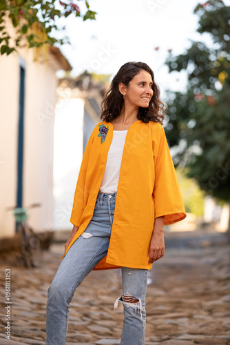 Young woman stands on a cobblestone street wearing a bright orange jacket with a white shirt and jeans during daytime in a quaint village