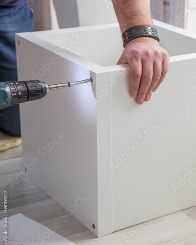 Man assembling white cabinet with cordless drill, driving screw into furniture panel, DIY home improvement, flat pack furniture assembly, handyman work, modern interior, close-up detail of