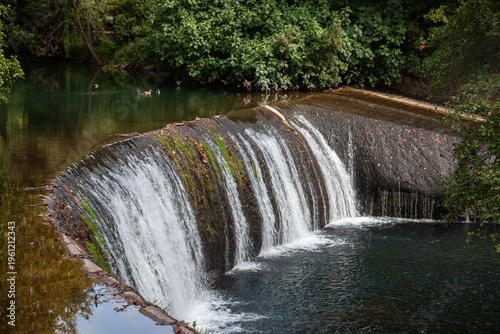 Schilderij op canvas Cascade Barrage Gasperini in Domaine de la Castille, La Crau, France, showing water flowing over a low dam into a calm basin surrounded by dense green vegetation