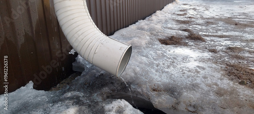 Fotografija Water trickles from a white plastic downspout onto a patch of melting, dirty snow and ice