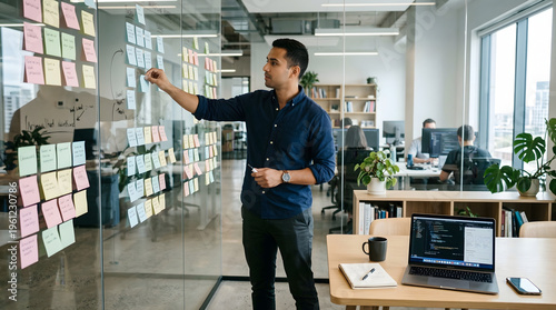 young professional male developer or project manager organizing colorful sticky notes on a glass wall in a modern open plan office during a creative brainstorming session