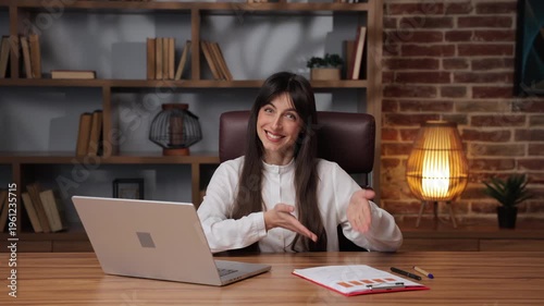 Business woman writes document and smiles in her office setting with laptop and books around.