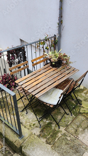 Small outdoor terrace with wooden table, folding chairs and potted plant near white wall. Cozy urban patio scene suitable for lifestyle content, home decoration ideas and small space design concepts.