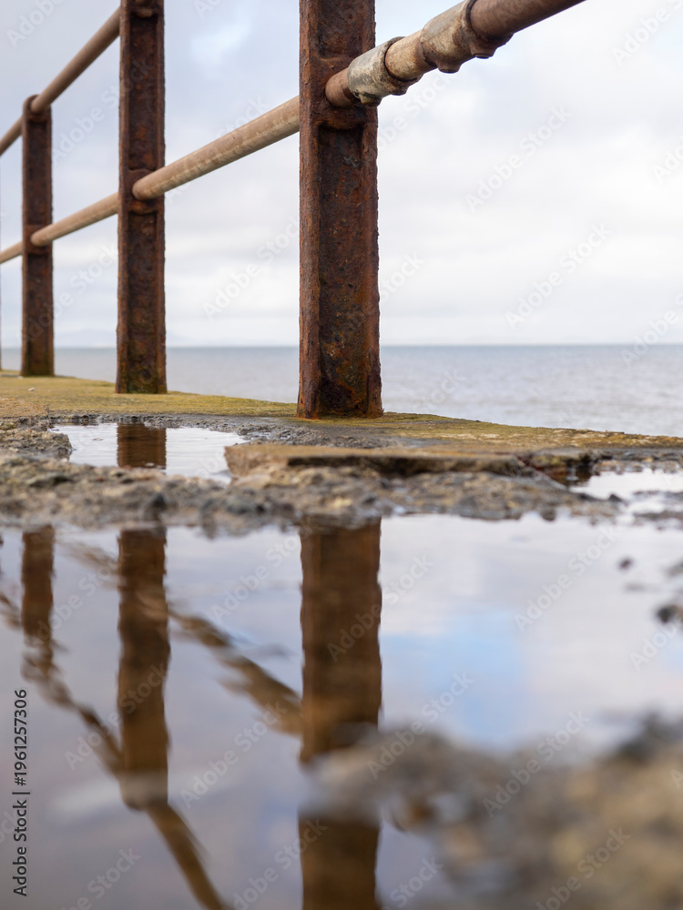 Fototapeta premium Rusty railing along the coast with reflections in puddles.