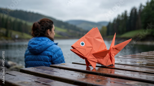 A woman sits on a wooden dock with an origami fish, overlooking a serene lake surrounded by trees and hills under a cloudy sky.