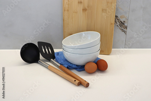 Kitchen utensils with bowls, wooden spatula, ladle and eggs cutting board on table. Simple cooking setup for food preparation, home kitchen routine, culinary process and domestic lifestyle concepts.