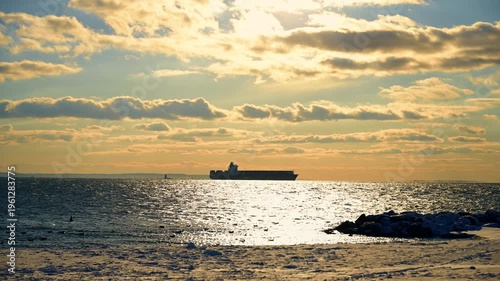 Seascape dazzled by the setting sun under the cloudy sky. Large freight boat stands on the roadstead in the horizon.