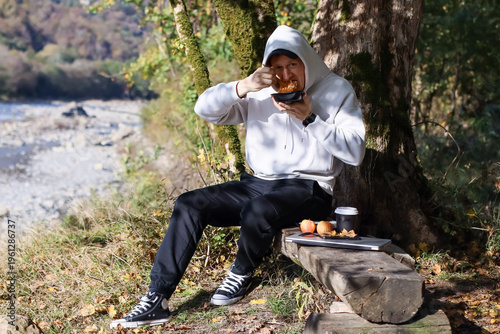 фотографія Hooded man eating snack with fingers by riverside