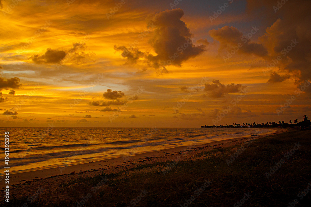 Fototapeta premium Golden sunset over ocean coastline with dramatic clouds and distant palm silhouettes