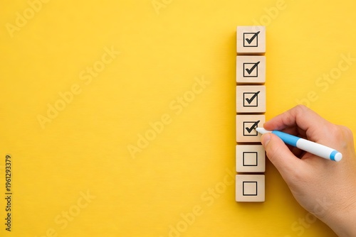 Hand with a blue pen is marking check boxes on a stack of wooden blocks against a bright yellow background, representing completion and achievement.