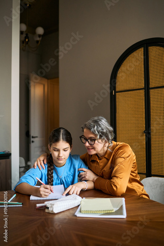 Vertical shot of middle aged woman guiding teenage girl writing in notebook at home, supporting homework and learning. Useful for education, parenting, tutoring, mentoring concepts