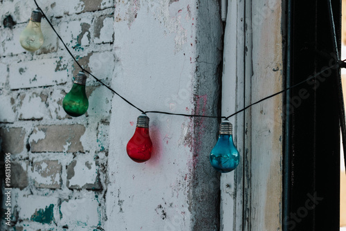 The image features a string of colorful light bulbs stretching across a weathered white brick wall. The green, red, and blue bulbs bring liveliness and a sharp contrast to the greyish urban environmen