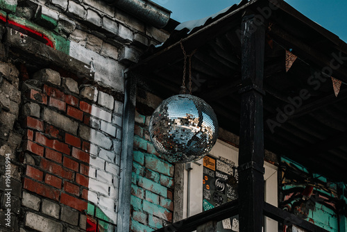 The image features a glittering disco ball hanging in an industrial setting against a weathered brick wall and wooden structures. Graffiti fragments and faded wall tones create a powerful contrast wit